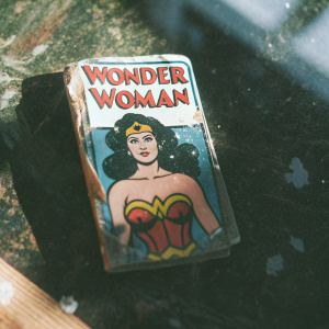 Vintage comic book card featuring Wonder Woman. Distressed and crumpled. Heroic pose, determined expression. Dark background, vibrant colors including red, yellow, brown, blue, and white. Stylized text in bold font.