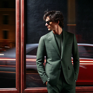 Christian Camargo, actor, with tousled dark hair, in a stylish olive green suit, blurred cityscape backdrop.