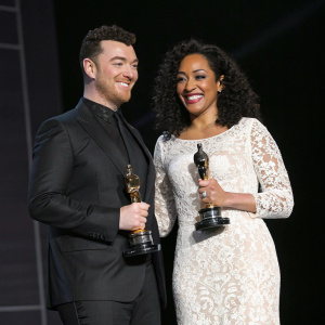 Sam Smith and Kanya King MBE standing on a stage, both holding awards. Sam in a sleek black suit, Kanya in a white lace gown. Against a dark, minimalist backdrop. Beaming smiles