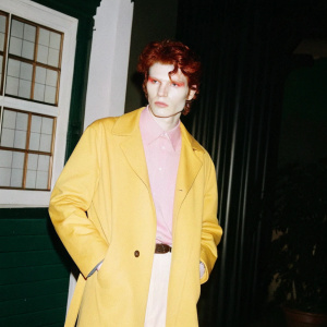 Gambit, a comic book character, depicted in vibrant graphic style. Red tousled hair, broad face, stern look, red eyes. Wears a long, colorful trench coat over a pink shirt.