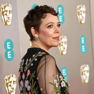 Close-up of Olivia Colman with short dark hair. Wearing a black top with floral embroidery and semi-transparent mesh fabric. Posing against a backdrop with the Hollywood News logo and EE emblem-like logos.