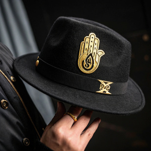 Close-up photo of a hand holding a black fedora hat from the hit anime series JoJo's Bizarre Adventure. The hat features a gold hand emblem prominently visible against a dark backdrop.