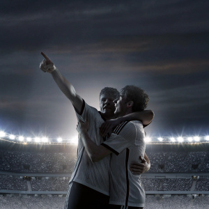 Toni Kroos and Leon Goretzka, soccer stars, celebrating victory with a passionate hug on the field. Toni Kroos points towards the horizon with a determined look. The backdrop is a sea of cheering fans and bright stadium lights.