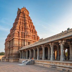A stunning photograph of the Sree Virupaksha Temple in Hampi, Karnataka, India. The temple's grandeur is highlighted by its detailed stone carvings and vibrant reddish-brown hues, set against a clear blue sky. Immaculate pillars and a towering, tiered structure dominate the frame.