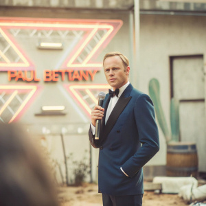 Paul Bettany, wearing a dark blue tuxedo, is at a press conference. Bold neon backdrop with event logos and text.