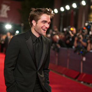 Robert Pattinson, dressed in a tailored black suit, grins broadly. Shiny red carpet backdrop, paparazzi flashes, blurred figures in the background