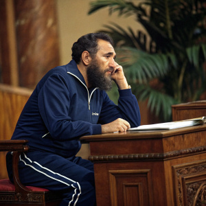 Fidel Castro, in a navy blue tracksuit, seated at a wooden desk. Deep in thought. Warm, earthy background