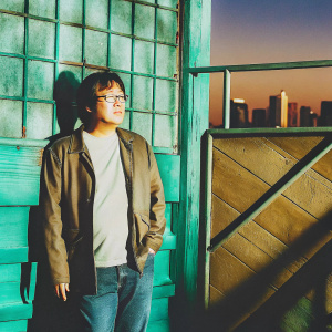 Masi Oka, Asian actor, in casual attire, posing outdoors at sunset. Golden hour lighting highlights his medium build, short dark hair, and glasses. A vibrant backdrop of a city skyline.