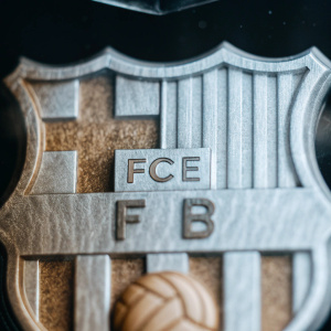 Digital grayscale image showcasing FC Barcelona badge. Shield-shaped design, textured and stitched, features patterned logo and 'FCE' text. Realistic embossed look with detailed lines and textures. Dark background.