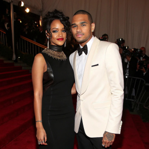 Chris Brown and Rihanna, both dressed formally, at a charity gala. Rihanna in a black dress, Chris in a tuxedo. The venue is dimly lit with red carpet and photographers in the background.