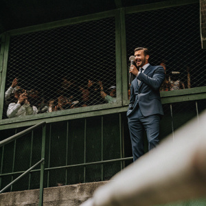 David Beckham, dressed in a blue suit, giving an interview. Crowd of fans in the background.