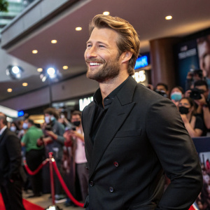 Glen Powell, with a beard and stylish hair, in a black suit, at the premiere of Top Gun: Maverick. Crowded theater lobby