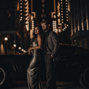 Vanessa Hudgens and Alex Pettyfer at the premiere event. Both in formal attire. She in a stunning silver gown, he in tailored suit. They pose confidently in front of neon lit cityscape backdrop.