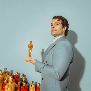 Henry Cavill, donning a tailored navy suit and curly hair, poses elegantly. Soft lighting, spotlight on him. Crowded awards ceremony backdrop