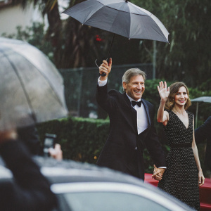 Harrison Ford and Calista Flockhart at the Golden Globe Awards. Ford in a classic black tuxedo, Flockhart in a black polka-dot dress. They are smiling and waving.