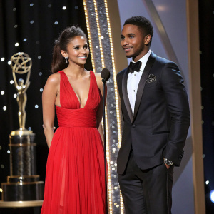 Nina Dobrev and Trey Songz, dressed in formal attire, at the Hollywood Film Awards. They stand beside the stage with the golden awards setup in the background.