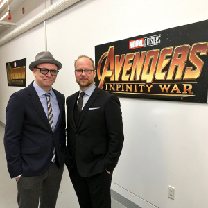 Christopher Markus and Stephen McFeely standing beside the Avengers: Infinity War banner. Both dressed in formal attire. Markus in a grey hat, glasses, dark blazer with striped tie. McFeely in a dark suit, solid tie, and a pocket square. They pose in a stark white room.