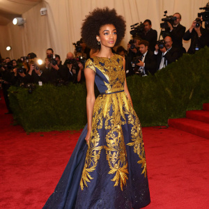 Esperanza Spalding in vibrant dress at Met Gala. Golden threadwork on dark blue fabric. Red carpet, paparazzi flashes.