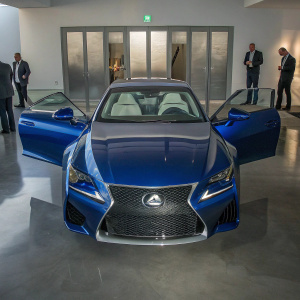A high-resolution image of a blue Lexus RC F in an upscale showroom. The RC F's sleek design and RCF badge prominently displayed. Interior details visible with doors open. Showroom featuring modern displays and attendees in business attire. Bright lighting emphasizes the car's glossy finish.