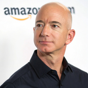 Professional headshot of Jeff Bezos, the visionary tech leader. Balding, serious expression, dark shirt. White backdrop with sleek Amazon logo