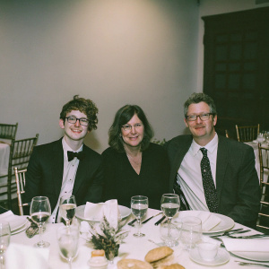 Tyler Oakley, Jackie Fields, and Ken Fields at a gala dinner. Tyler with a youthful vibe, Jackie with a warm smile and medium-length hair, Ken with a mature look. Formal attire, elegant table setting with wine glasses, plates, silverware, and bread rolls. Warm lighting, indoor setting.