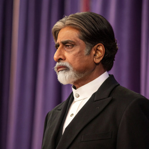 Amitabh Bachchan, looking solemn. Black suit, white shirt, traditional mustache and beard. Purple curtain backdrop.