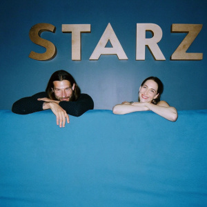 Zach McGowan and Emily Johnson at a promotional event. Smiling in front of black backdrop with golden STARZ logo.