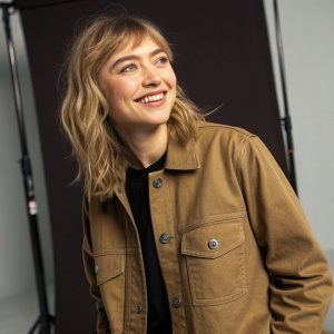 Imogen Poots wears the Carhartt-WIP Owen Shirt Jacket. She smiles warmly. The jacket features button closures and a pocket. Simple studio backdrop