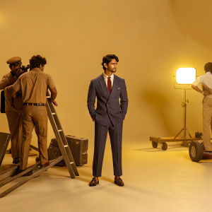 Sushant Singh Rajput, dapper in a navy blue suit with a red tie, graces the bustling set of a Bollywood film. Vibrant studio lights and crew members surround him.