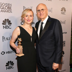 Kasia Ostlun and Jeffrey Tambor attend a glamorous awards ceremony. Kasia in a sleek black dress, Jeffrey in a tailored dark suit. They stand against a backdrop filled with various sponsor logos.