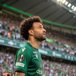 Felipe Anderson, wearing a green jersey with sponsors' logos, during a match at a packed stadium. Intense look