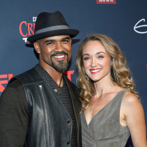 Shemar Moore and Lauren Wood at a glamorous gala. Shemar in a black fedora and leather vest, Lauren in a gray dress. Both smiling, posing close together. Dark blue backdrop with bold logos and elements. Close-up color photo.
