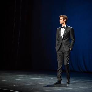 Andrew Garfield, in a sleek black suit and bow tie, standing alone on an empty stage. Dark blue backdrop