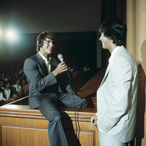 Carlos Pena and Logan Henderson, from Big Time Rush, in a candid chat during a press conference. Formal attire, stage setup with audience in background