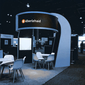 Eberlehaid's booth at the construction expo. Sleek design, curved display with company logo. Seating area with modern silhouettes. Color scheme of blue, white, gray, and brown.