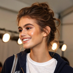 Close-up of Kaia Gerber at the New York Fashion Week. Model with warm-toned skin, joyful expression, casual outfit, and voluminous hair. Soft background lights.