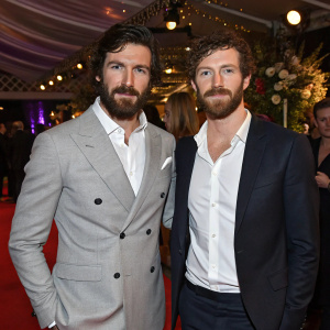 Eoin Macken and Nat Sanders at a glamorous event. Eoin in a stylish gray suit, Nat in a crisp white shirt. Both sport beards. Festive backdrop