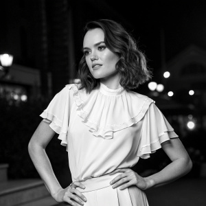 Caucasian actress Daisy Ridley in elegant black and white photoshoot. Standing confidently, medium length brown hair. Simple yet stylish ruffled blouse. Dark background with soft lighting