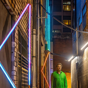 Dean Winchester, Supernatural character, in vibrant green shirt. Dark urban alley background, neon lights casting shadows