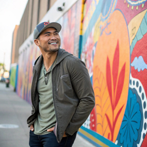 Martin Sensmeier, smiling confidently, in a casual outfit. Standing outdoors in front of a vibrant mural wall