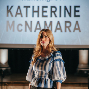 Katherine McNamara, blonde actress, wearing a blue and white striped blouse with ruffles, standing confidently on a stage with large, bold letters on a light-colored backdrop.