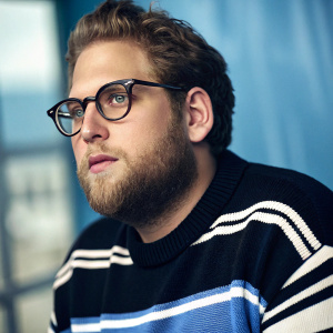 Close-up shot of Jonah Hill. Caucasian male with glasses and full beard. Wearing a black, white, and blue striped sweater. Intense gaze. Blurred blue background.