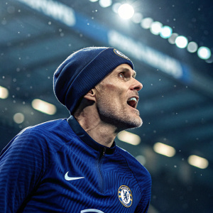 Thomas Tuchel, looking intense and emotional. Wearing blue Chelsea FC jersey and a beanie. Stadium lights blurred in the background.
