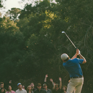 Jordan Spieth mid-swing at the PGA Tour event. Dressed in blue polo shirt, khaki pants. Crowd cheering in background.