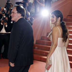 Quentin Tarantino and Daniela Pick at Cannes Film Festival. He's in a black suit, she's in a white dress. Surrounded by flashing cameras and reporters.
