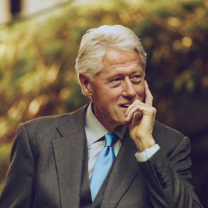 Former US President Bill Clinton in a thoughtful pose. Grey suit, light blue tie. Solo shot, blurred dark background