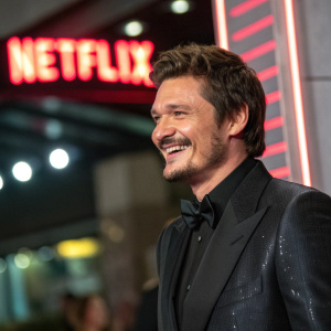 Pedro Pascal in a sleek black suit, smiling confidently. Close-up portrait. Brightly lit, bold neon Netflix logo in background.
