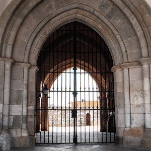 A grand archway leading to the Abbey of Santa Maria la Real de Las Huelgas in Burgos, Spain. Constructed from large, weathered stones with a classic arch design. A sturdy black iron gate frames the central opening, contrasting against the lighter stone background. The gate, with dark iron bars, provides a peek into the abbey's architecture. The focus is on the stone masonry and the timeless, robust aesthetic typical of traditional European styles.
