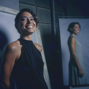Close-up studio portrait of Aubrey Plaza. Wearing a sleek black sleeveless dress and a warm smile. White background with minimal text elements. Emphasizes upper body