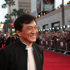 Jackie Chan, smiling warmly, at the Hollywood premiere. Wearing a red carpet outfit. Large crowd in background